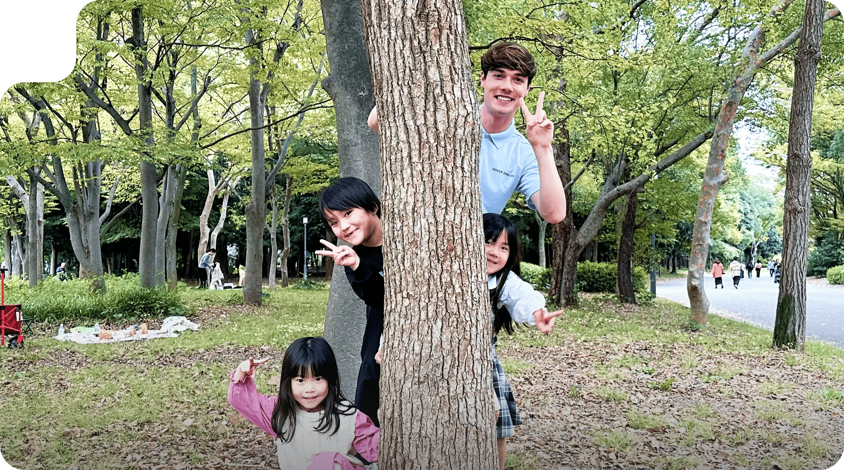 子どもたちと講師が木の陰から顔を出しこちらに笑顔を向けています